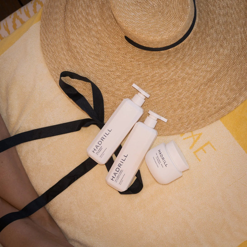 Hadrill The Shampoo, The Conditioner and The Treatment Mask on a yellow beach towel beside a straw hat